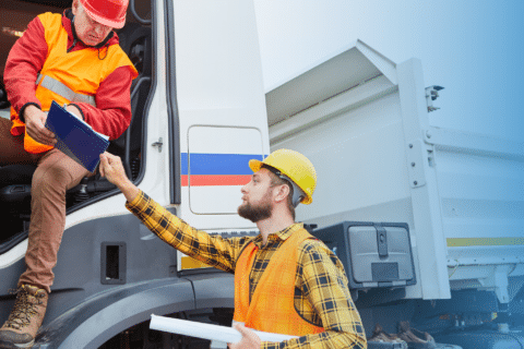 Two construction workers in safety gear are adjacent to a truck. One sits in the drivers seat, holding a clipboard, exemplifying key strategies for fleet managers, while the other stands outside, reaching for documents.