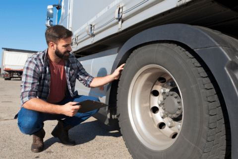 A person kneels next to a large truck, inspecting the front tire with a clipboard during a CVSA Roadcheck, on a paved outdoor area.