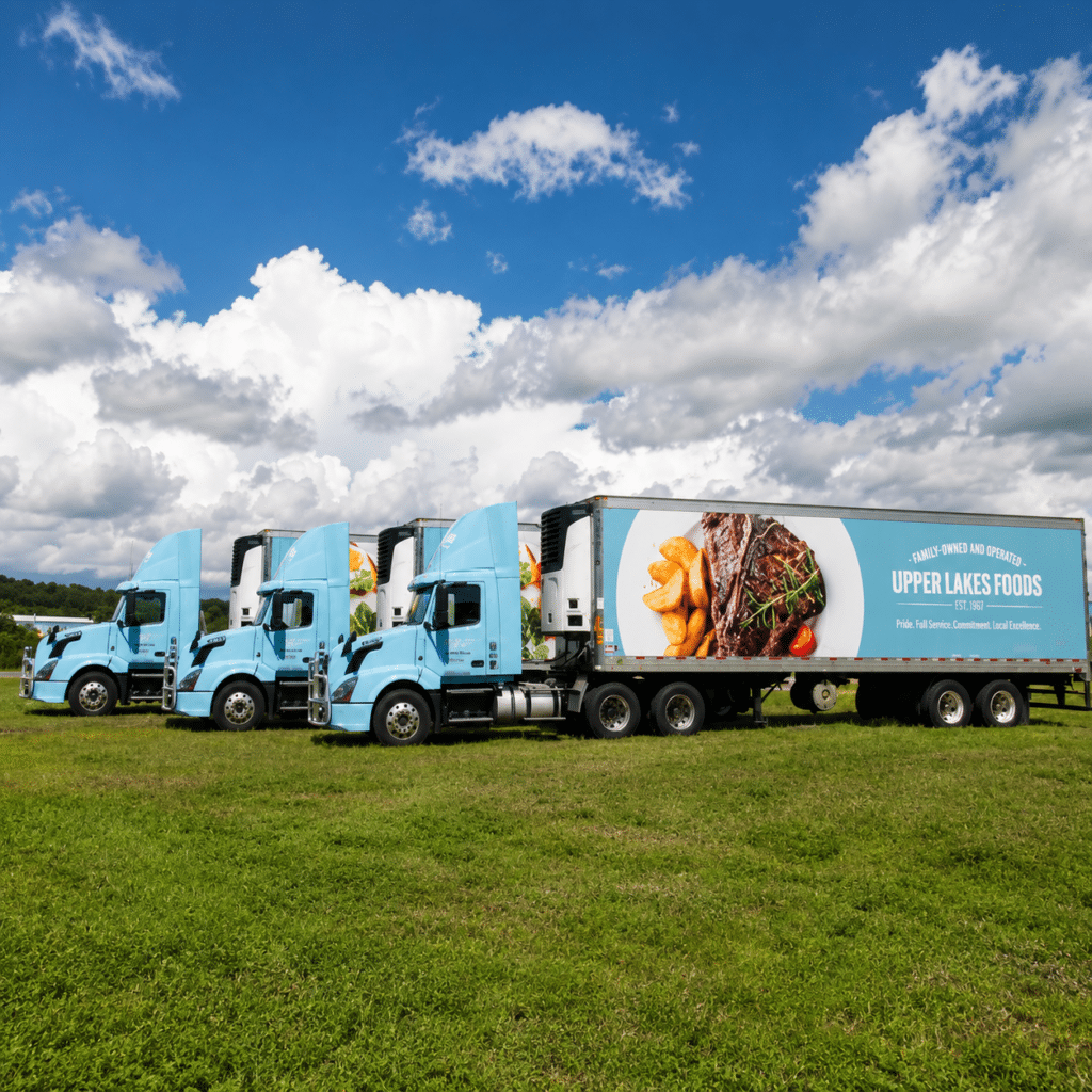 Four light blue Upper Lakes Foods delivery trucks with food-themed advertisements are parked on grass under a partly cloudy sky.