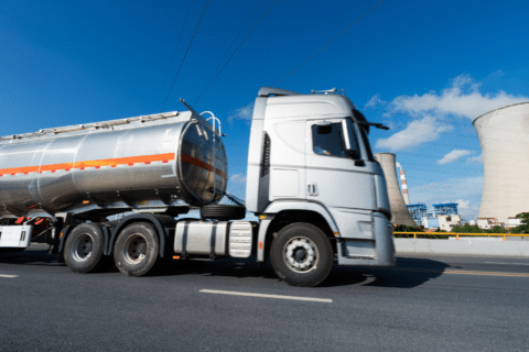 A silver tanker truck drives on a road with large cooling towers and industrial buildings in the background under a blue sky.