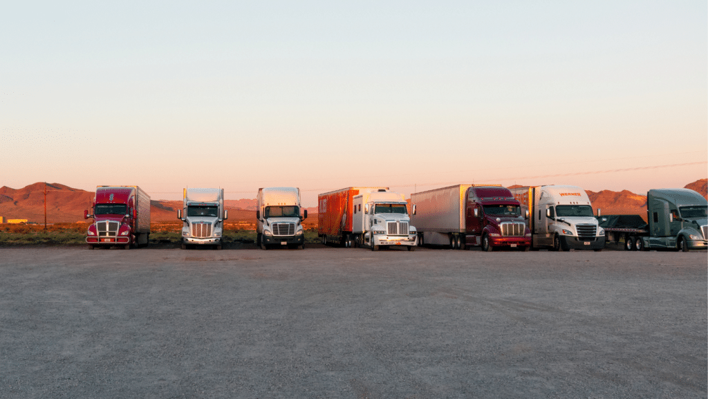 Six semi-trucks, ideal for various CDL Classes, are parked side by side in a gravel lot, backed by mountains and a clear sunset sky.